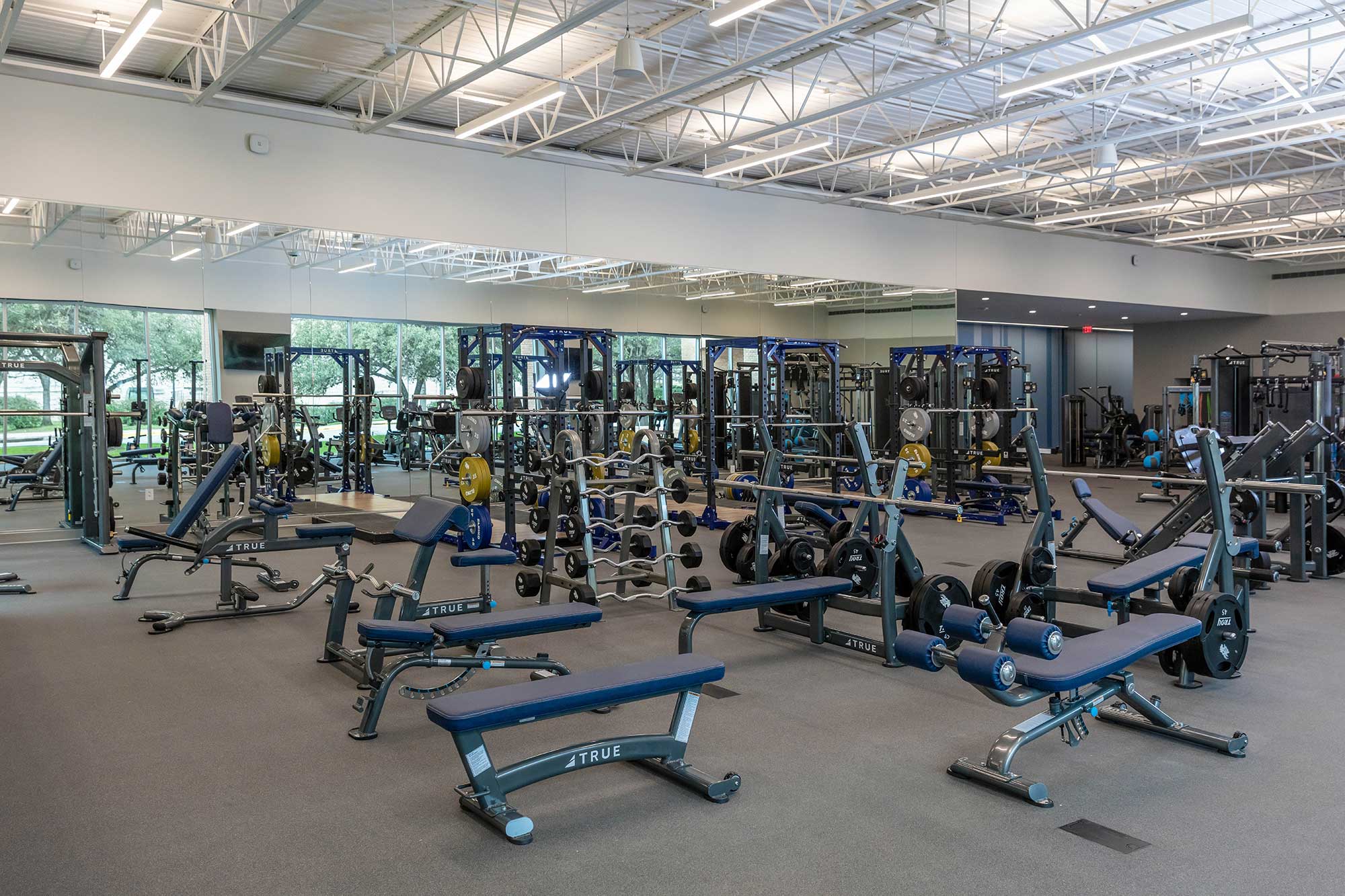 Spacious gym with multiple weight benches, squat racks, weight plates, dumbbells, and exercise machines. Large mirrors line one wall, and windows provide natural light. The gym is clean and empty.