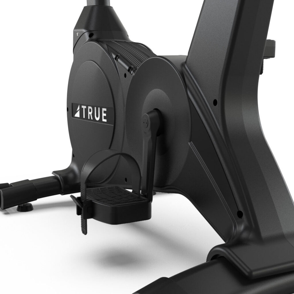 Close-up view of the pedal and flywheel area of a black stationary exercise bike, featuring the brand name “TRUE” on the side panel.