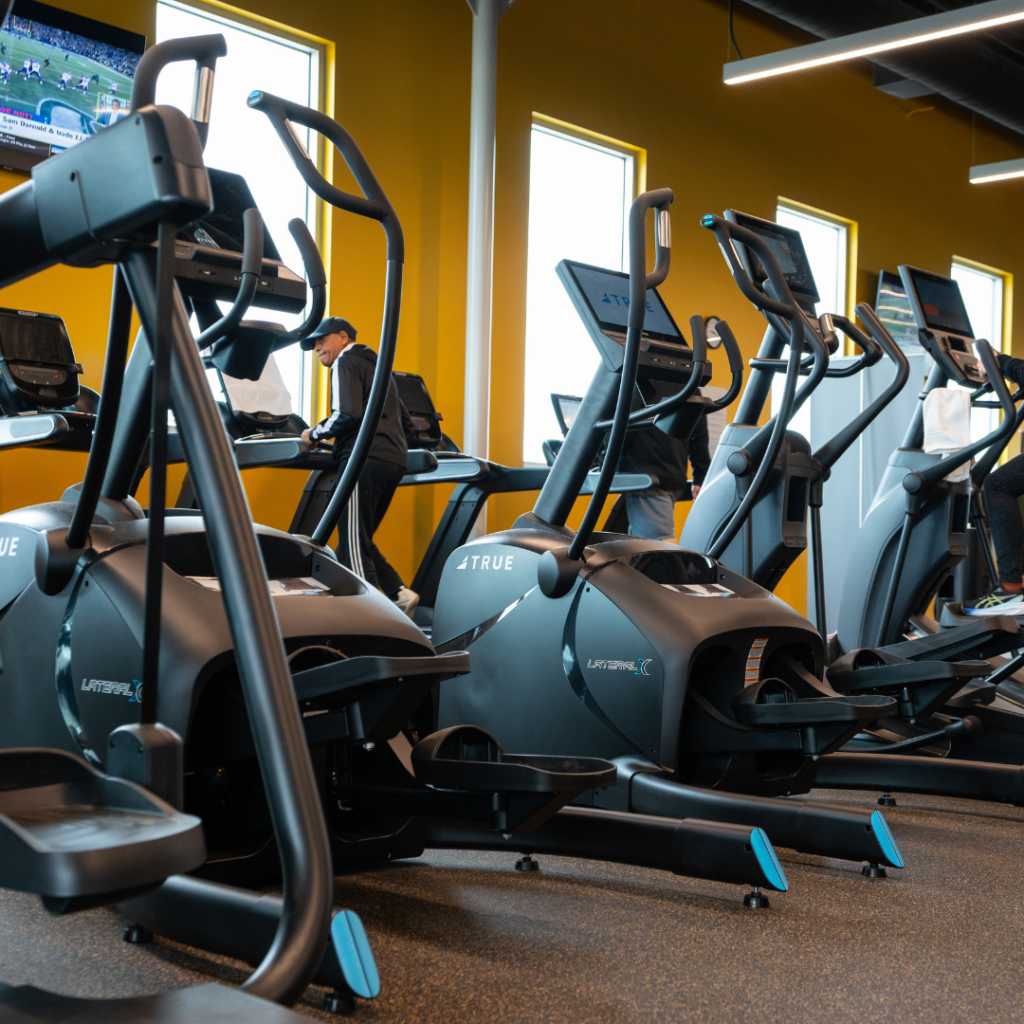 A row of elliptical machines in a gym with yellow walls, large windows, and people exercising. A TV mounted on the wall displays a sports game.
