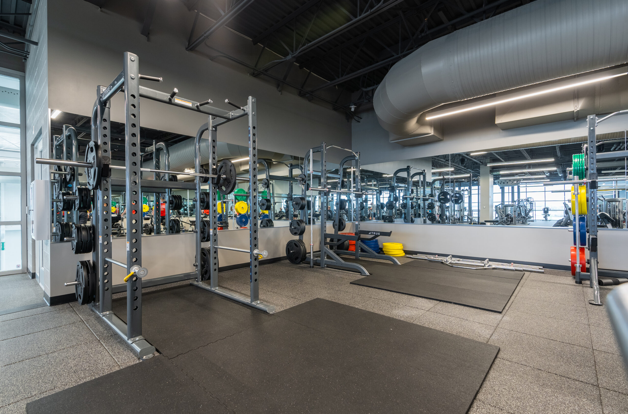A modern gym with weightlifting equipment, including squat racks, barbells, benches, and mirrors lining the walls. The space is clean, well-lit, and has rubber flooring.
