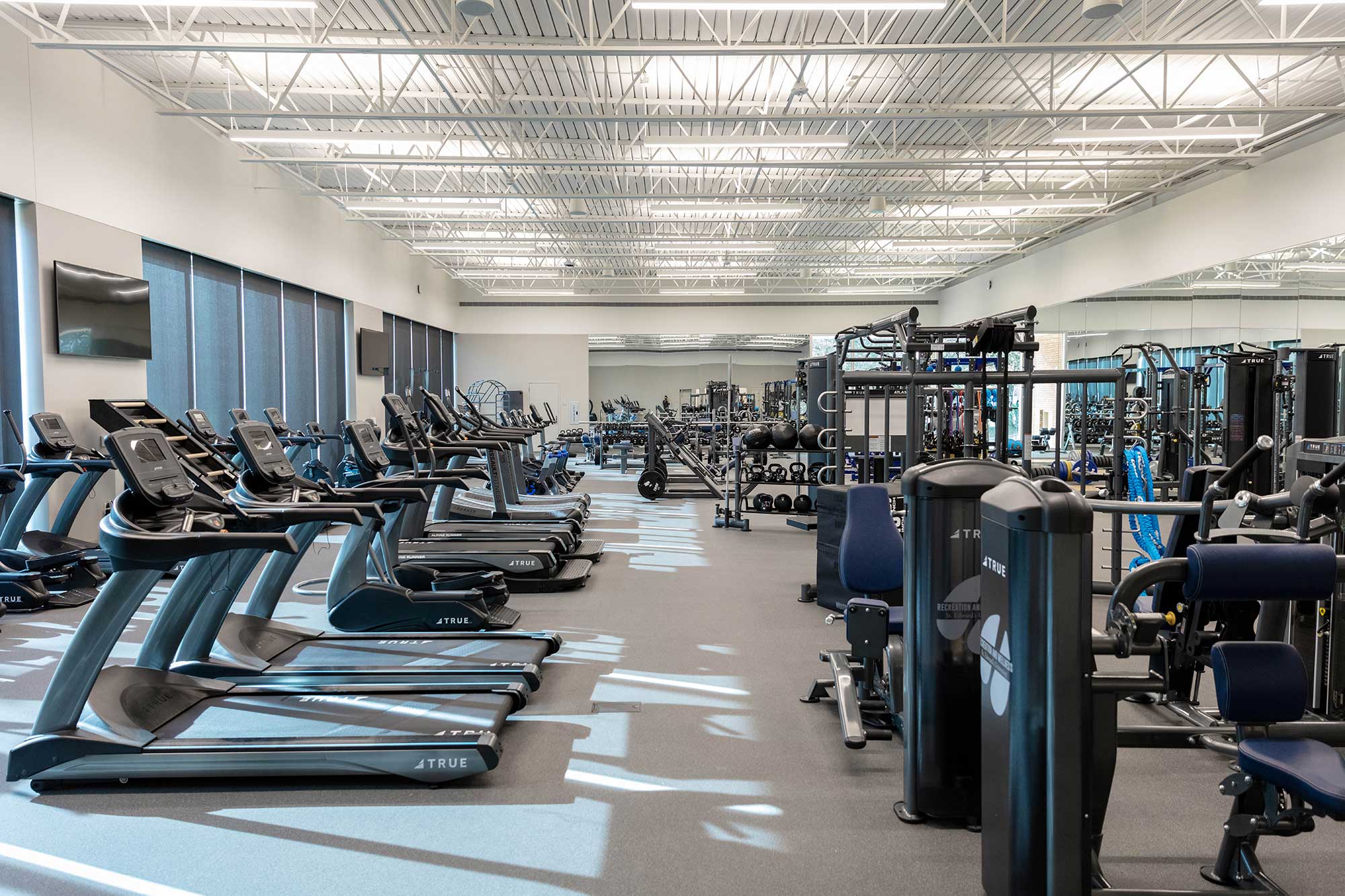 Spacious gym with rows of treadmills, weight machines, and free weights. Large windows line one wall, letting in natural light. The gym is clean, organized, and empty of people.