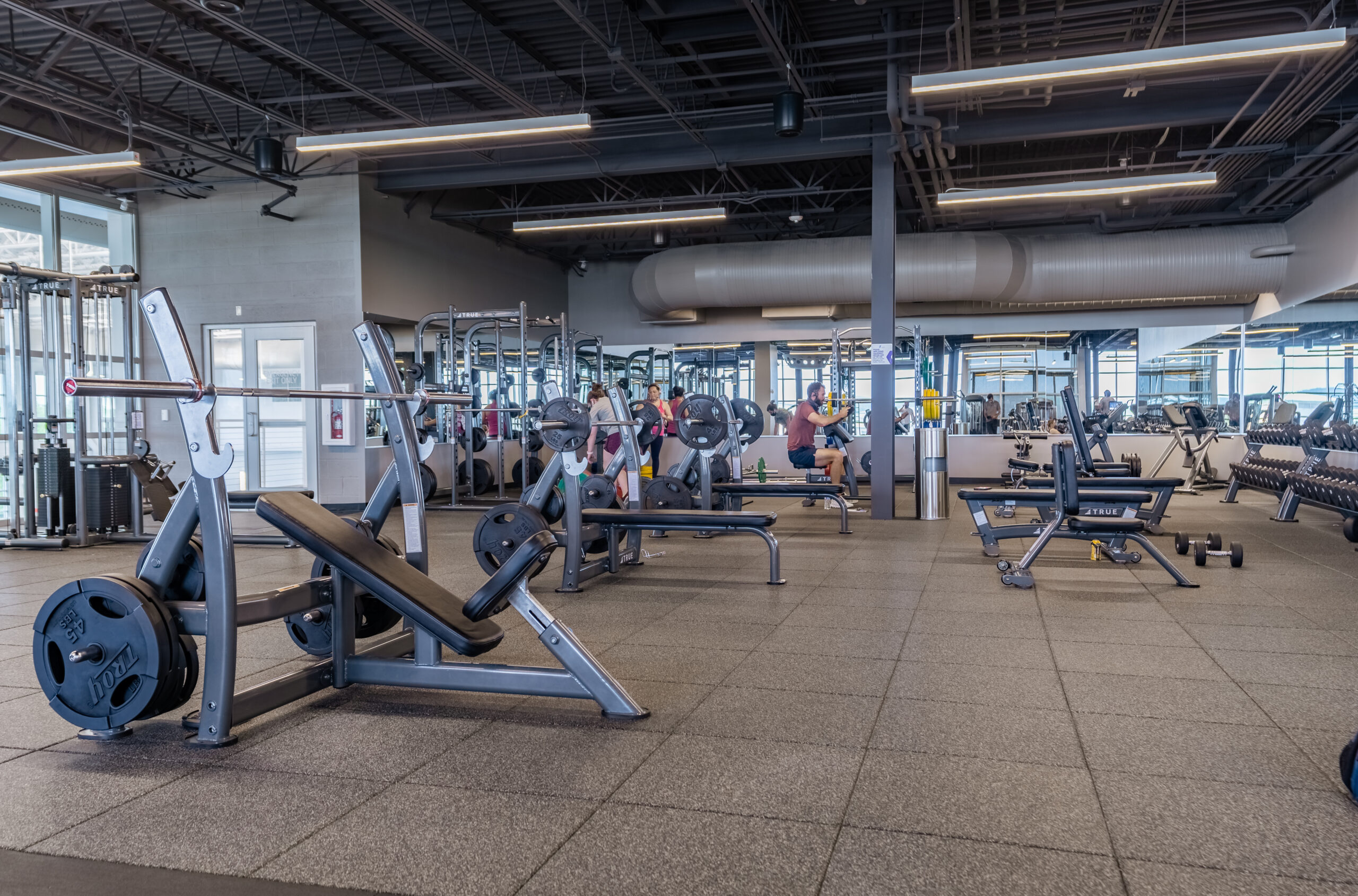 Large gym with weightlifting equipment, benches, and free weights on a rubber floor. Few people are working out in the background near mirrors and racks. The space is spacious and well-lit with industrial ceilings.