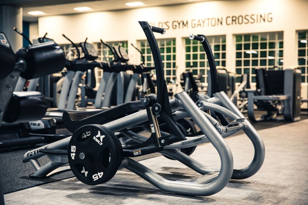 A modern gym with various exercise machines and free weights. In the foreground, plate-loaded gym equipment is neatly arranged. The gym appears bright and spacious with large windows in the background.