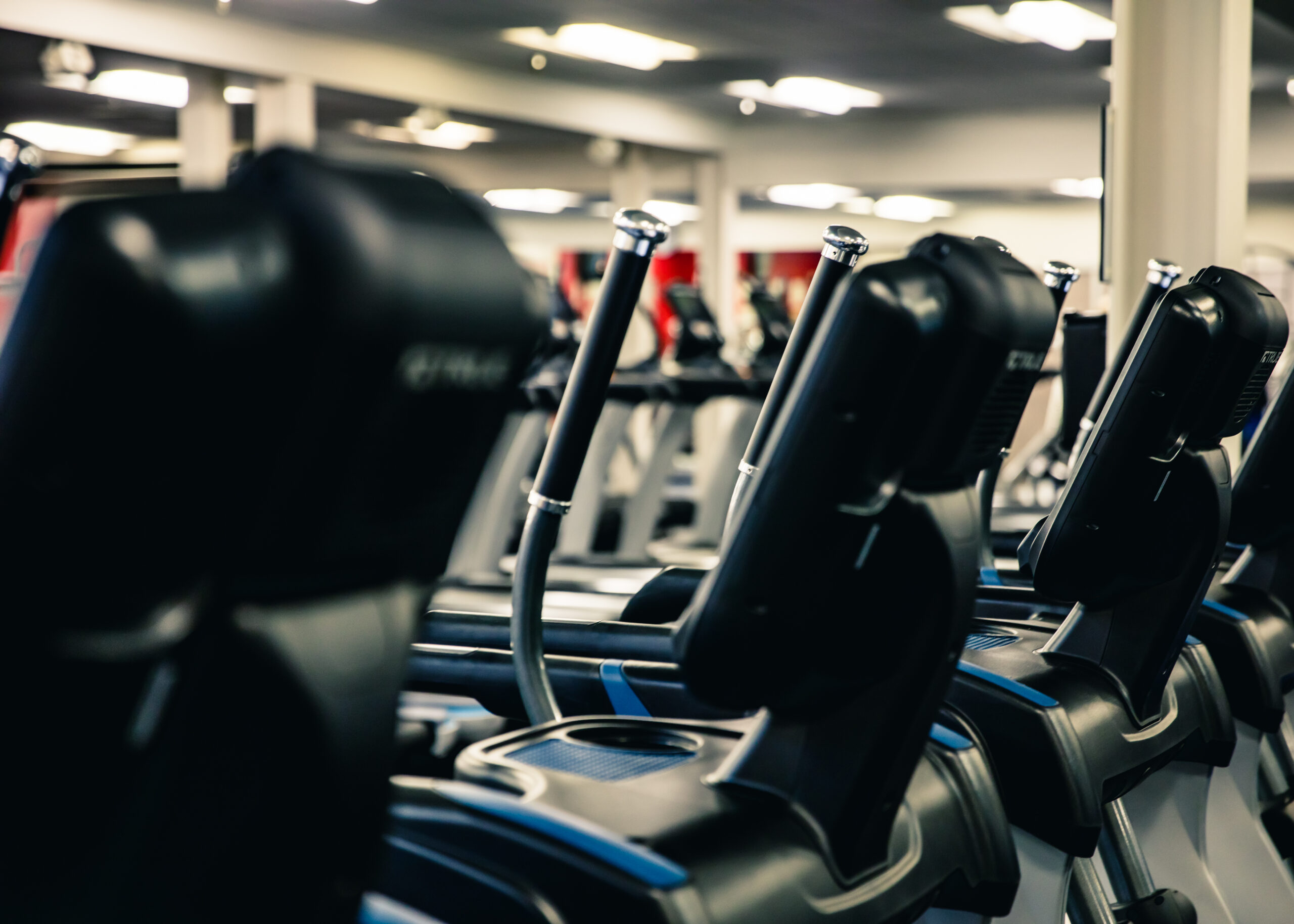 Row of elliptical machines in a brightly lit gym, focused on the handlebars and control panels, with more fitness equipment visible in the blurred background.