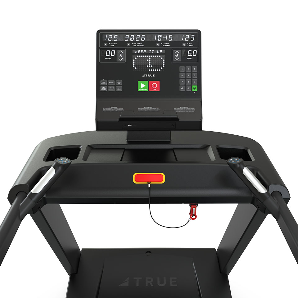 Close-up of a treadmill console displaying workout data including time, distance, calories, and speed, with buttons for starting, stopping, and adjusting settings. A safety key with a red clip is attached below the console.