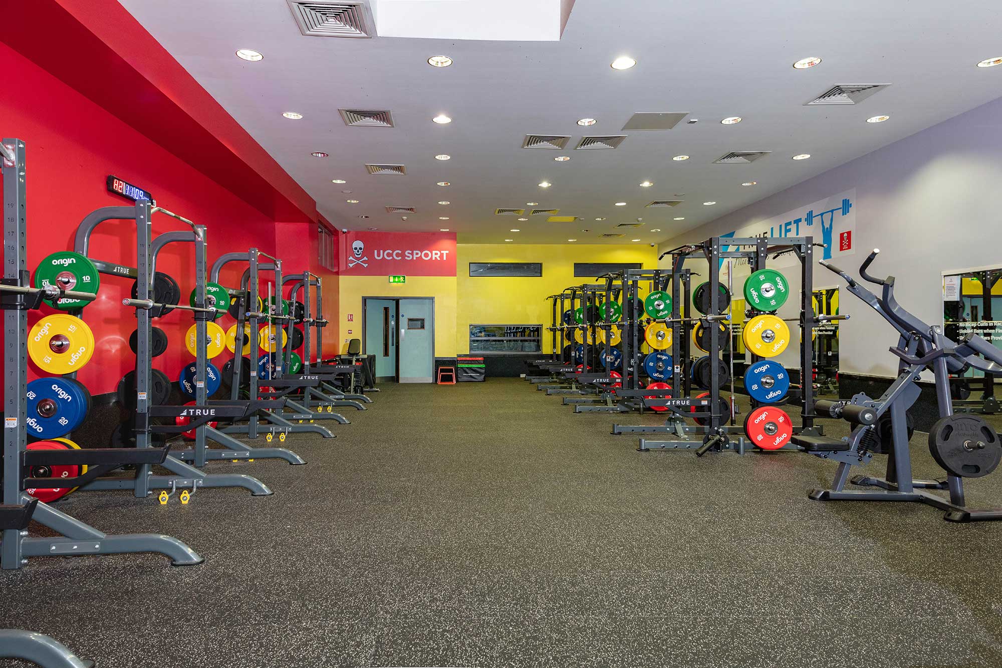 A spacious gym with weightlifting racks, colorful weight plates, and various fitness equipment. The walls are painted red and yellow, and a sign reads UCC SPORT near the back entrance. The gym is well-lit and empty.