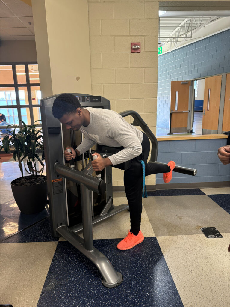 A man in a white shirt and bright orange shoes uses a leg curl machine at a gym, lifting one leg behind him while gripping the handles and looking downward.