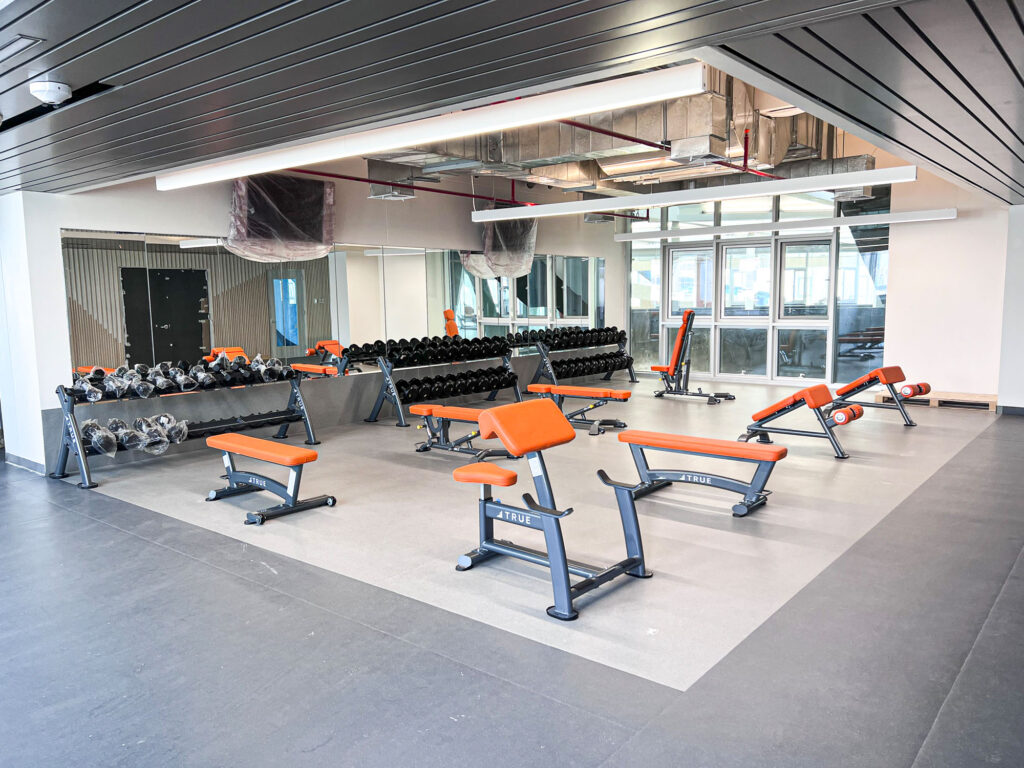 A modern gym with orange and gray weight benches, dumbbells on racks, and exercise equipment. Large mirrors cover one wall, and windows let in natural light. The room has a clean, open, and organized layout.