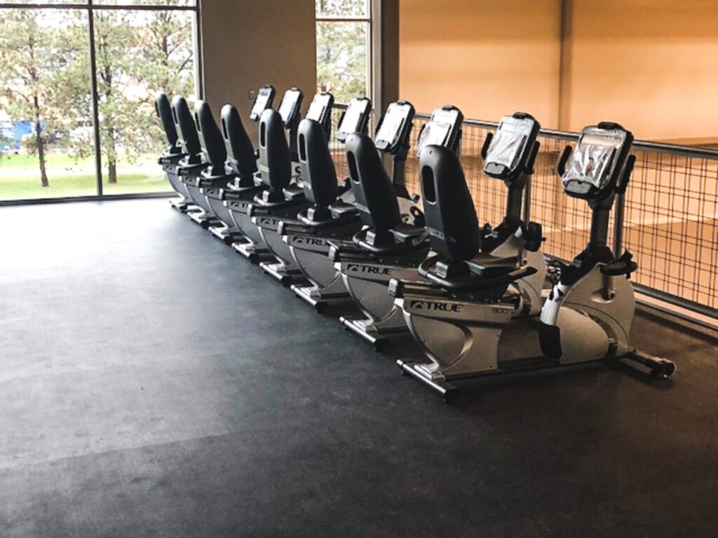 A row of stationary exercise bikes lined up in a gym with large windows, allowing natural light to enter and providing a view of trees outside. The gym floor is dark and spacious.