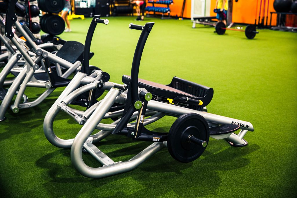 A leg press exercise machine with weights is positioned on green artificial turf inside a brightly lit gym with orange walls and various workout equipment in the background.