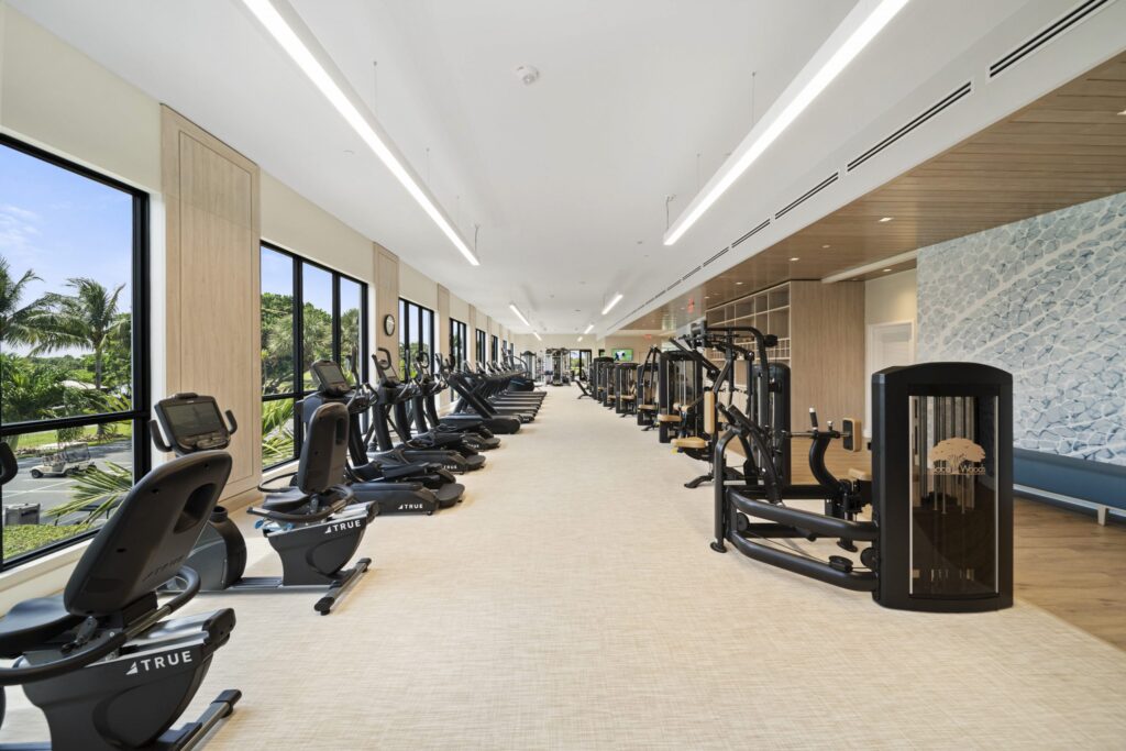 Spacious modern gym with large windows, several cardio and strength training machines, light-colored flooring, and bright natural light coming in. Trees and blue sky are visible outside.