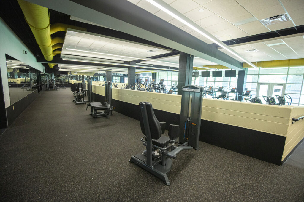 Spacious gym with exercise machines, including leg extension equipment in the foreground, large mirrors on the left wall, and rows of cardio equipment near large windows in the background.