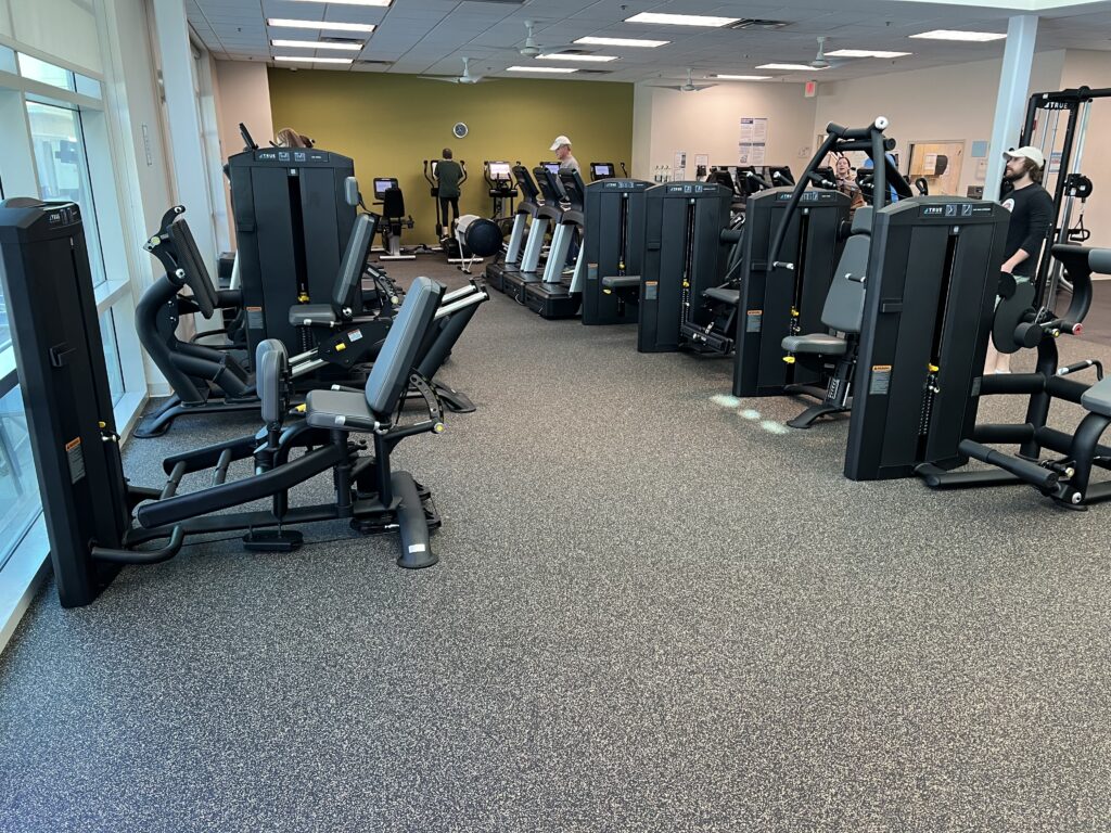 A spacious gym with various black exercise machines, including leg and arm equipment, arranged on a speckled floor. A few people are working out, and large windows let in natural light.