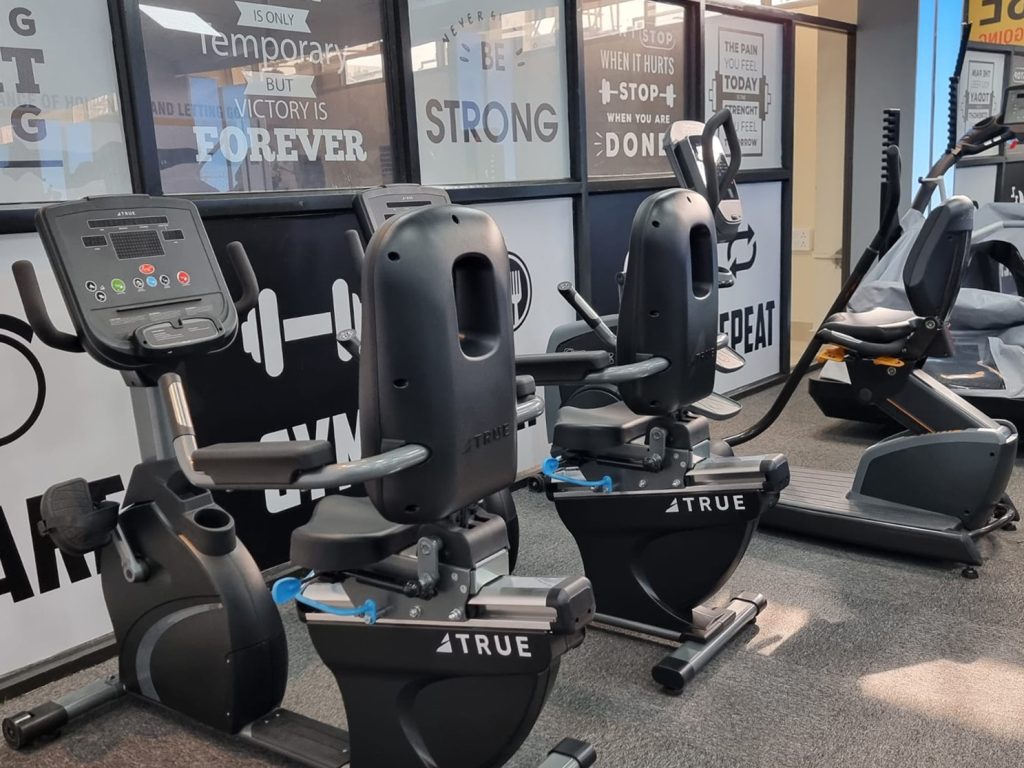 Two stationary exercise bikes and an elliptical machine are positioned in a bright gym, with motivational quotes and fitness-related images decorating the wall behind them.