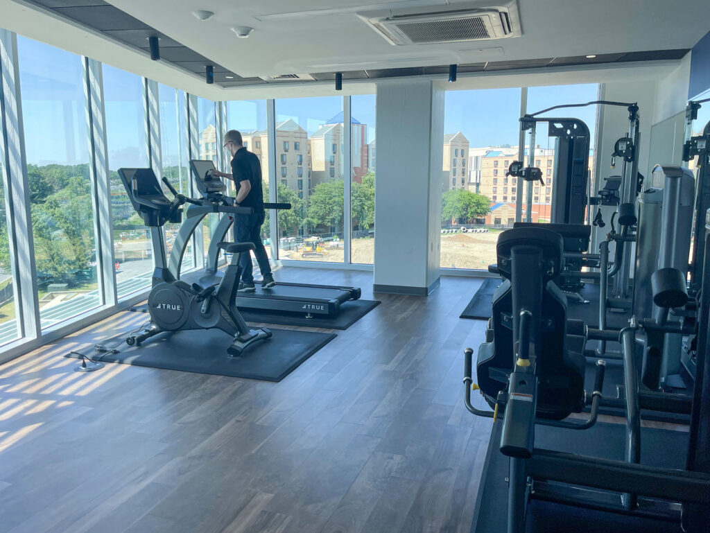 A person uses a treadmill in a modern gym with large windows, letting in natural light. Various exercise machines are visible, and buildings and trees can be seen outside.