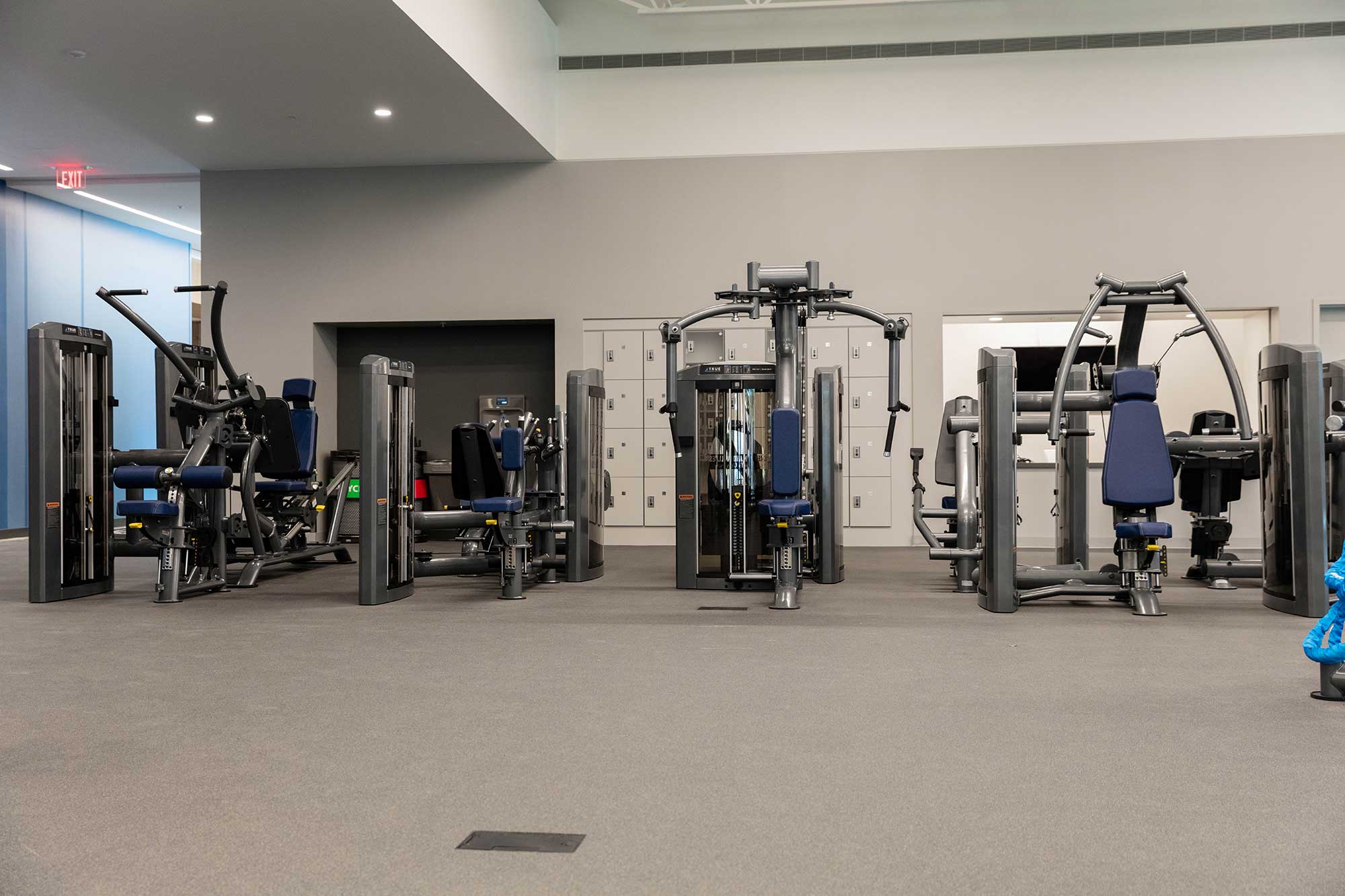 A spacious gym with various strength training machines on a carpeted floor, blue cushioned seats, and lockers in the background. The area is clean and well-lit, with no people present.