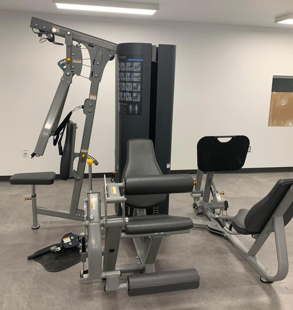 A leg extension and curl machine, along with a leg press machine, are seen in a clean, modern gym with gray flooring and white walls. Both machines are unoccupied and placed near each other.