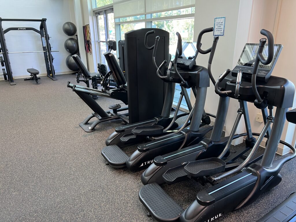 A gym room with ellipticals, a seated leg press machine, an exercise bike, stability balls, and a large strength training station, all on rubber flooring near large windows.