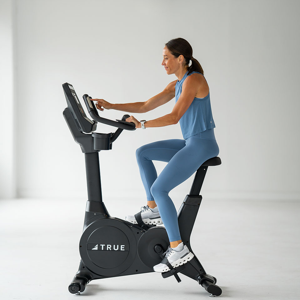 A woman in a blue workout outfit exercises on a stationary bike indoors, smiling as she uses the touchscreen display. The bike is branded TRUE and the room has a clean, minimalistic look.