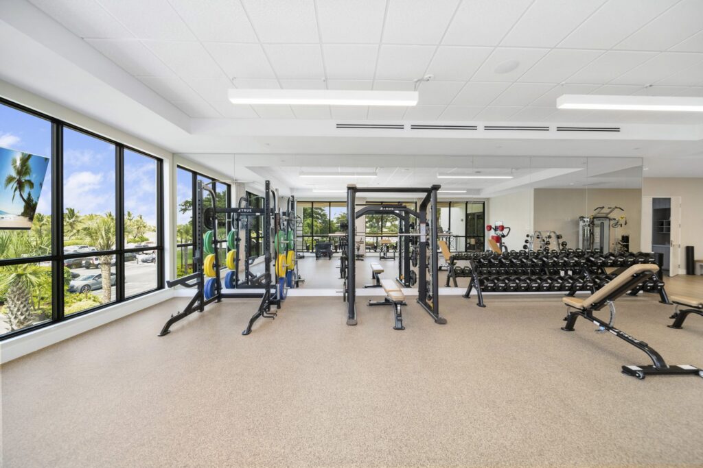 A bright, spacious gym with large windows, weightlifting machines, benches, and a rack of dumbbells. Palm trees and blue sky are visible outside. The room has beige flooring and a white ceiling with fluorescent lights.