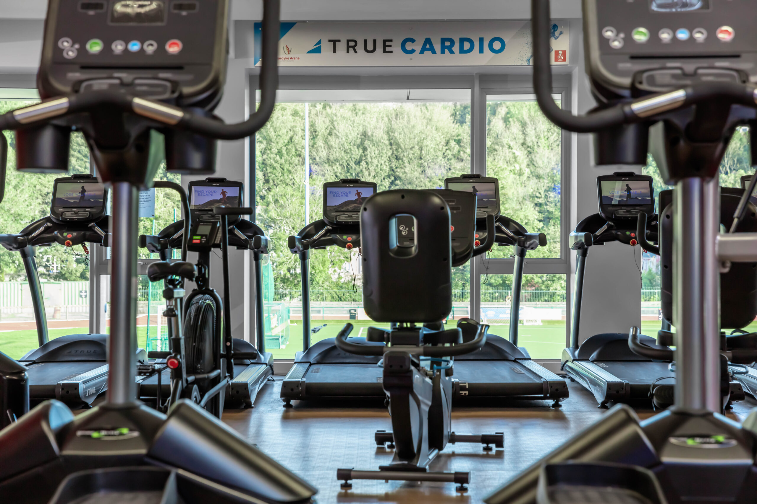 A row of treadmills and exercise bikes face large windows in a bright gym with the words “True Cardio” displayed on the wall above, and greenery visible outside.