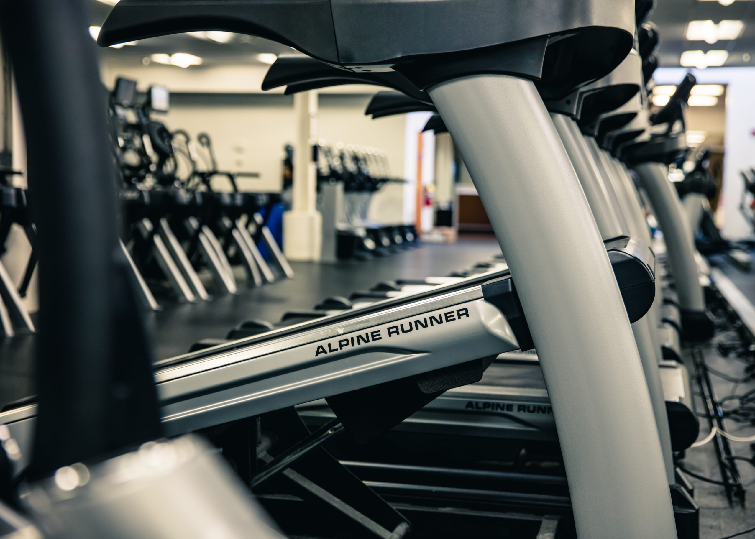 Close-up view of several treadmills labeled Alpine Runner lined up in a gym, with other exercise equipment visible in the background.