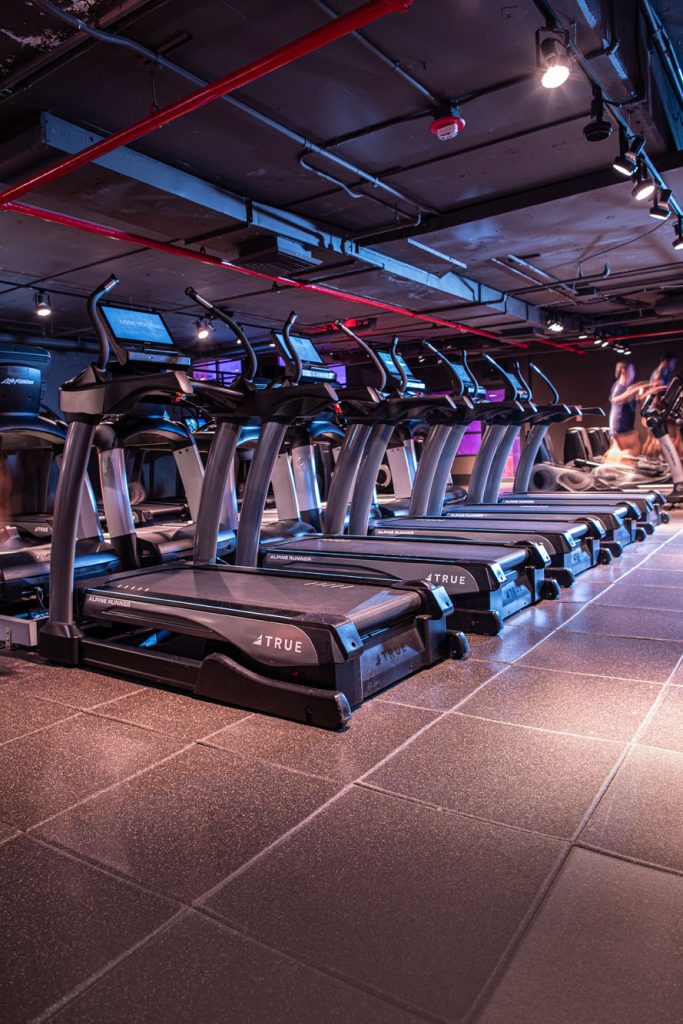 A row of treadmills in a modern gym, illuminated by overhead lights, with a few people working out in the background. The space has a dark ceiling with red pipes and tiled flooring.