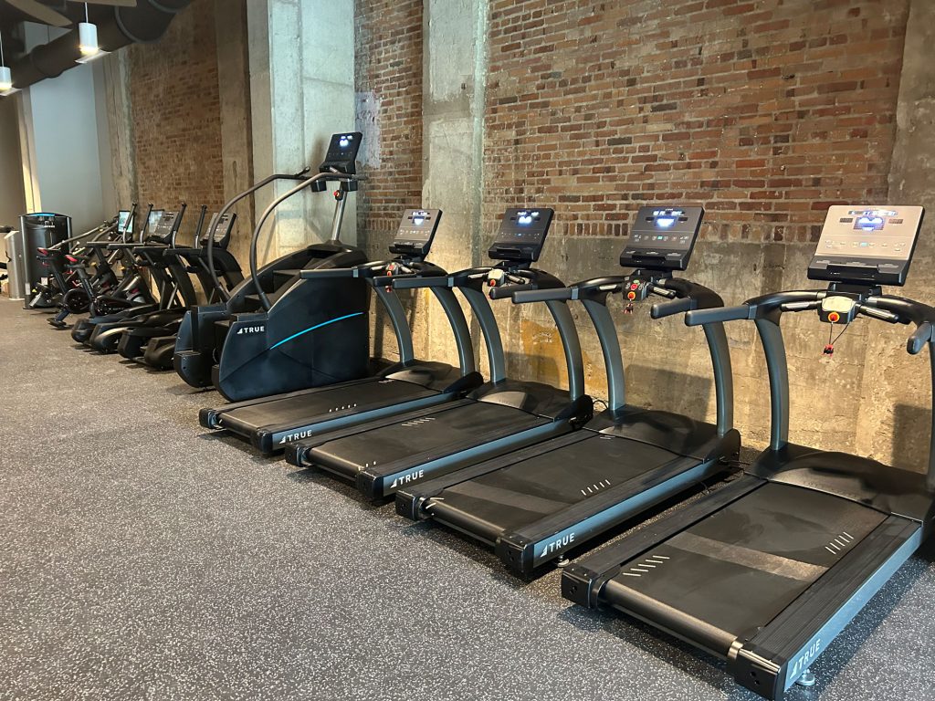A row of treadmills and elliptical machines lines a gym with exposed brick walls and rubber flooring, all equipment turned off and neatly arranged.