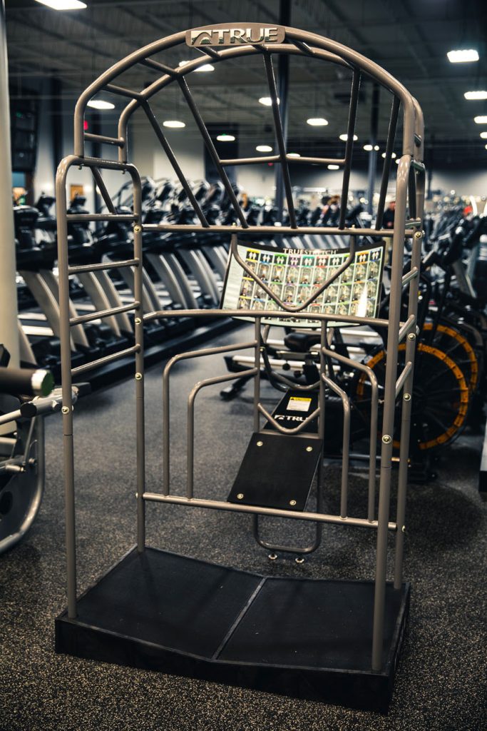 A standing calf raise exercise machine in a gym, with instructional diagrams on a panel and various other exercise equipment visible in the background.