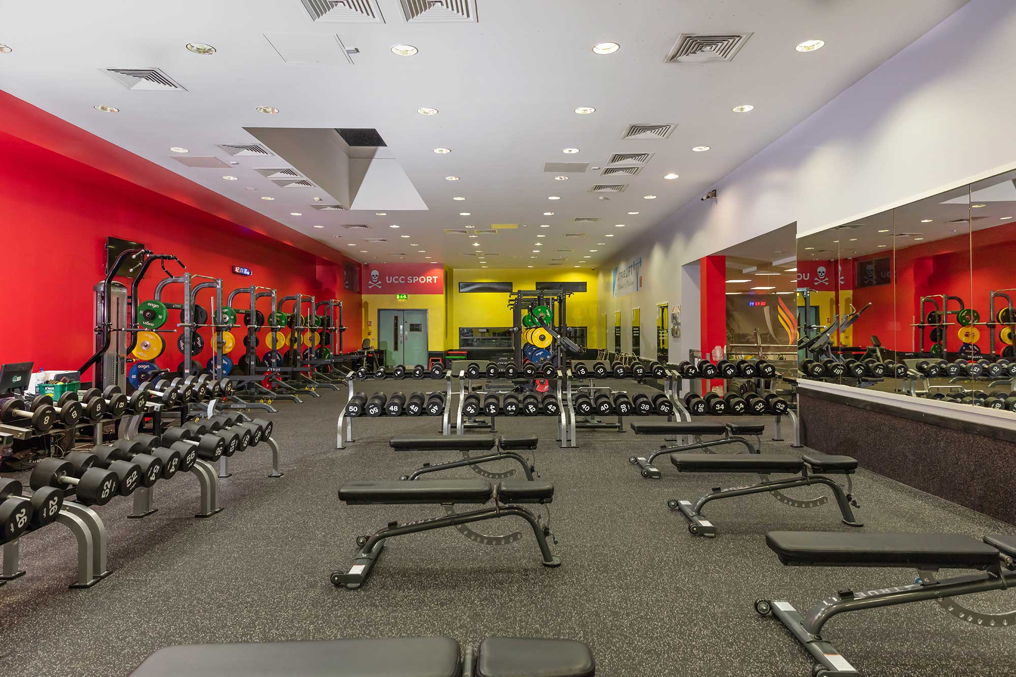 A spacious gym with red and yellow walls, lined with free weights, benches, barbells, and weight racks. Large mirrors cover one wall, reflecting the equipment and bright overhead lighting.