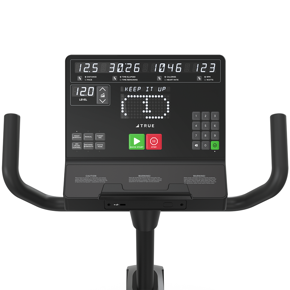 Close-up of a stationary bike display showing workout data such as time, distance, speed, and calories. The screen displays KEEP IT UP with handlebars visible on either side.