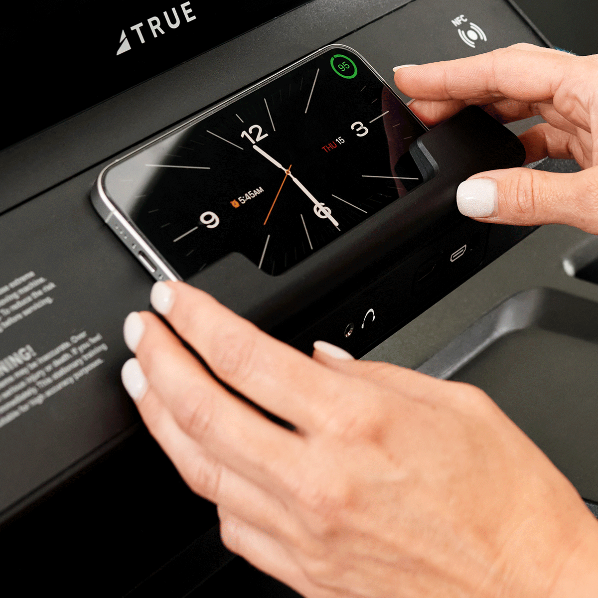 A person places a smartphone displaying a clock screen onto a fitness equipment console with wireless charging and control features.