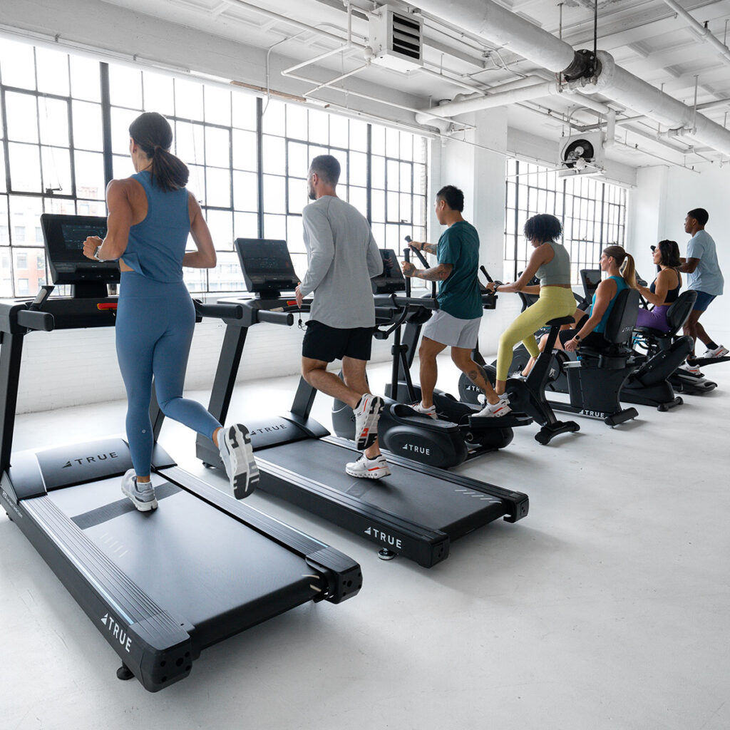 A group of people exercise indoors, using treadmills and stationary bikes lined up by large windows in a bright, modern gym.