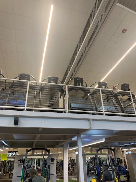 A gym interior with a mezzanine level holding treadmills, seen from below. The lower level features various fitness equipment and weights. Bright lights and a high ceiling illuminate the space.