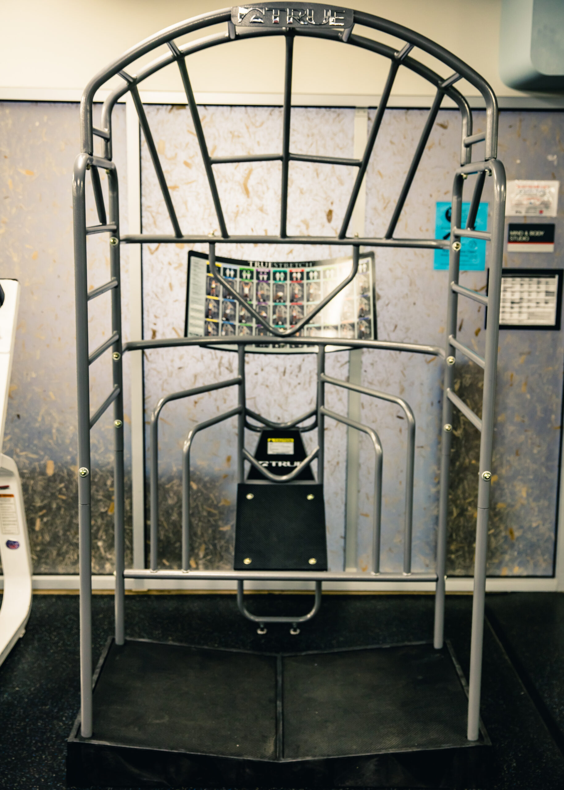 A machine with a metal frame and padded seat, designed for stretching or flexibility exercises, stands in a gym with gray walls and informational posters in the background.