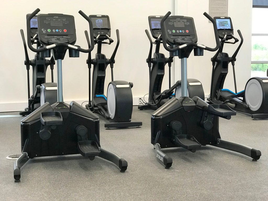A gym with several black elliptical machines and two recumbent stepper exercise machines on a gray floor, positioned in front of a white wall with windows to the right letting in natural light.