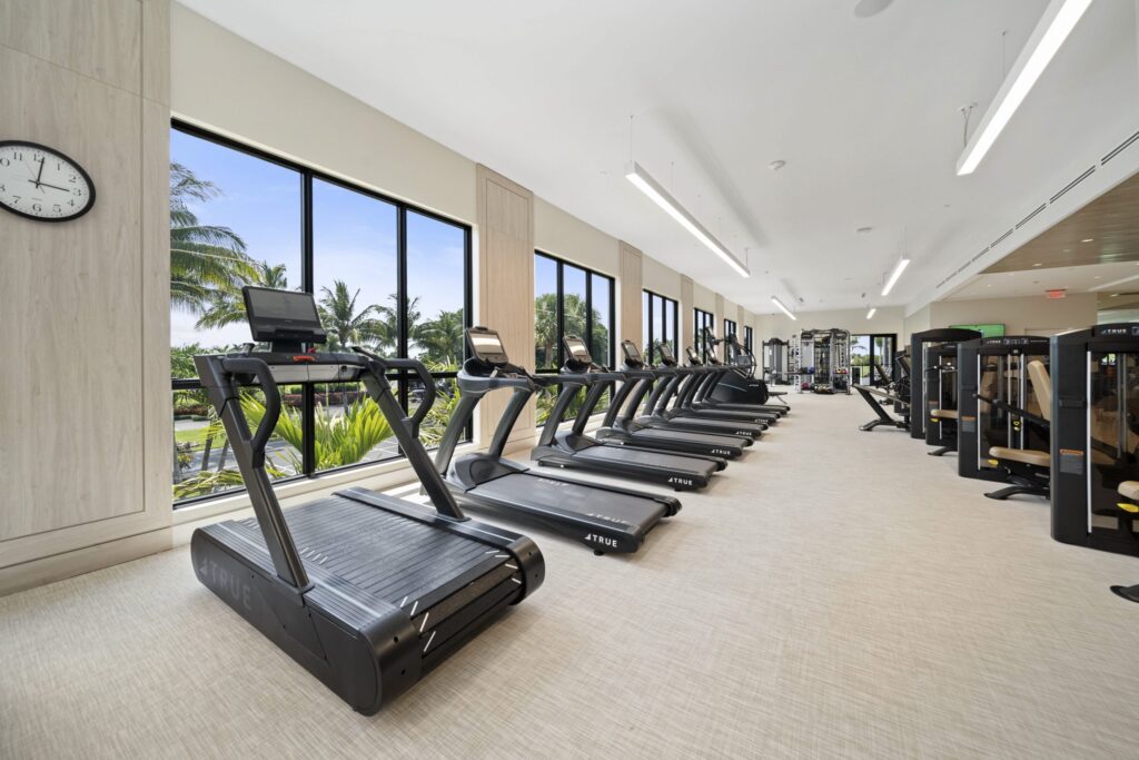 A bright, spacious gym features several treadmills lined up beside large windows with views of palm trees and greenery. The room is clean, modern, and well-lit with exercise machines in the background.