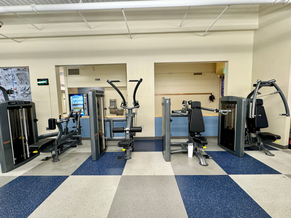 Three strength training machines are lined up in a gym with blue and gray checkered flooring; a water dispenser and lockers are visible in the background.