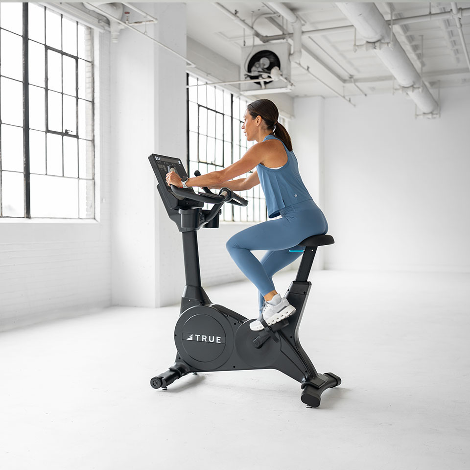 A woman in a blue workout outfit rides a stationary exercise bike in a bright, spacious gym with large windows and white walls.
