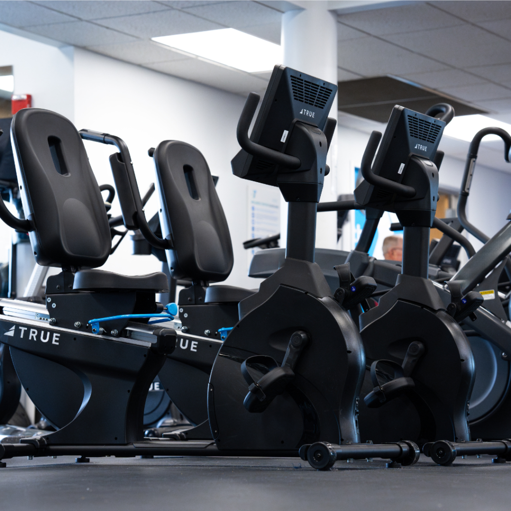 A row of black stationary exercise bikes with digital screens is arranged in a bright, modern gym with white walls and ceiling panels.