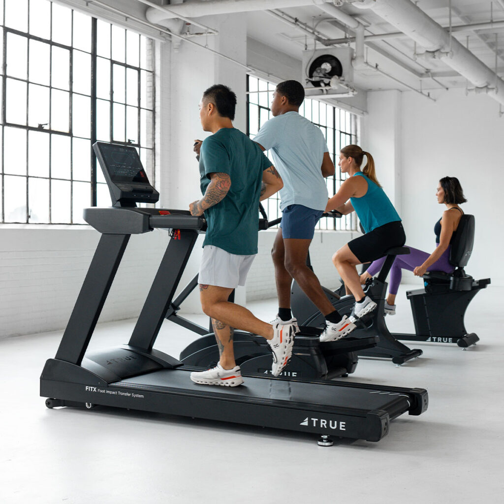 Three people exercise on treadmills while one person uses a stationary bike in a bright, spacious gym with large windows and white walls. All are wearing athletic clothing and focused on their workouts.