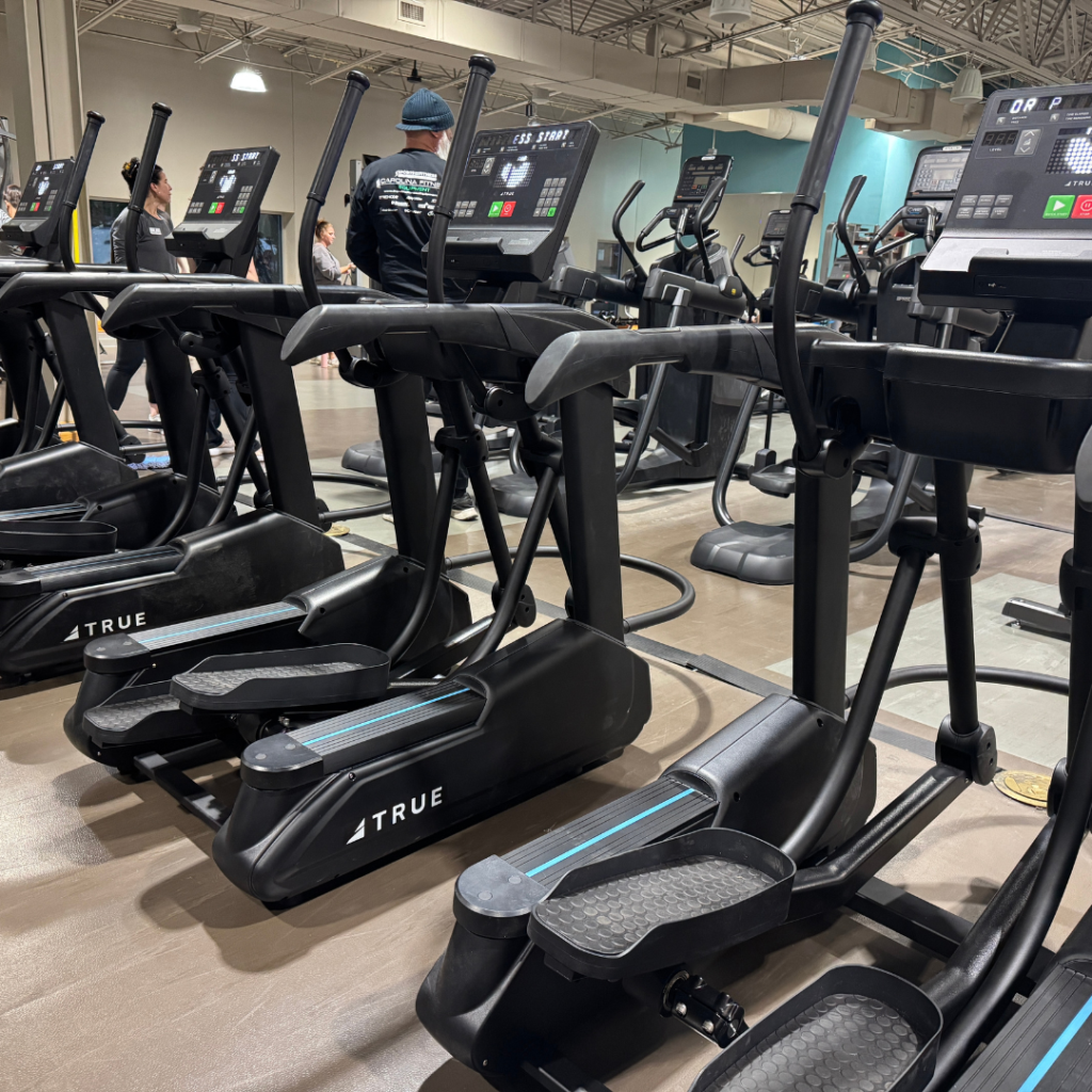 A row of black TRUE elliptical machines is set up in a gym. The gym has high ceilings, overhead lighting, and several people working out in the background.