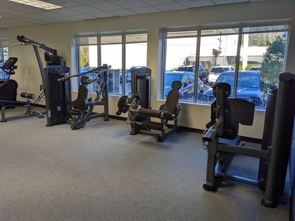 A gym room with various strength training machines positioned near large windows; outside, several cars are parked and trees are visible through the windows. The room is well-lit and has a clean, spacious feel.