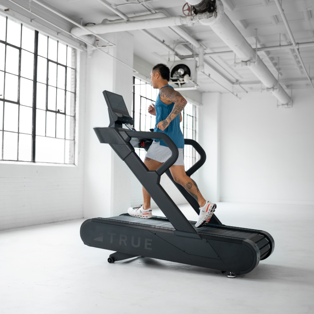 A person with tattoos runs on a treadmill in a bright, spacious gym with large windows and white walls. The treadmill is black and modern, and the runner wears a blue tank top and white sneakers.