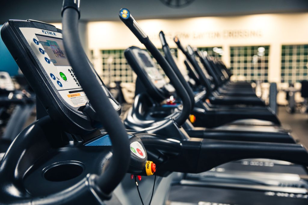A row of modern treadmills in a brightly lit gym, with digital displays and control panels visible in the foreground. The background is slightly blurred, highlighting the exercise equipment.