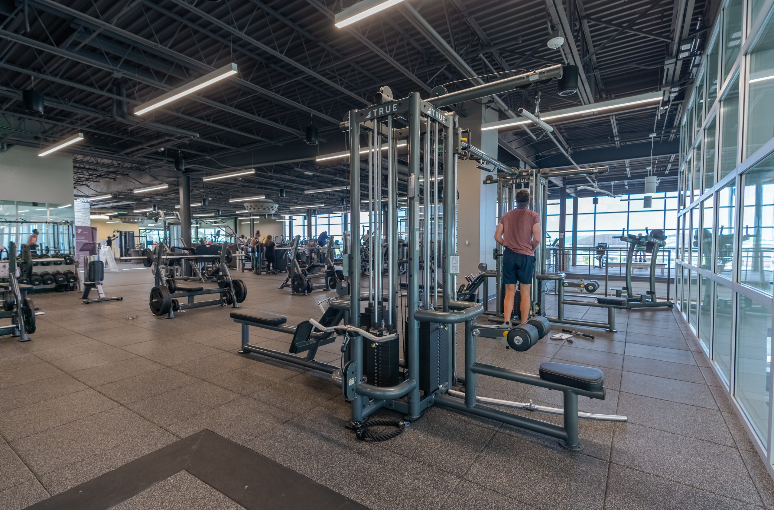A spacious gym with various exercise machines and equipment, including weightlifting stations. Several people are working out, and one person in a pink shirt is using a cable machine near large windows.