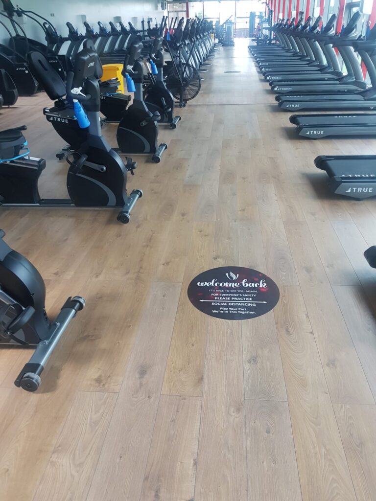 A gym with rows of treadmills on the right and exercise bikes on the left. A circular floor sticker in the center reads “welcome back.” The gym has wooden floors and large windows at the far end.