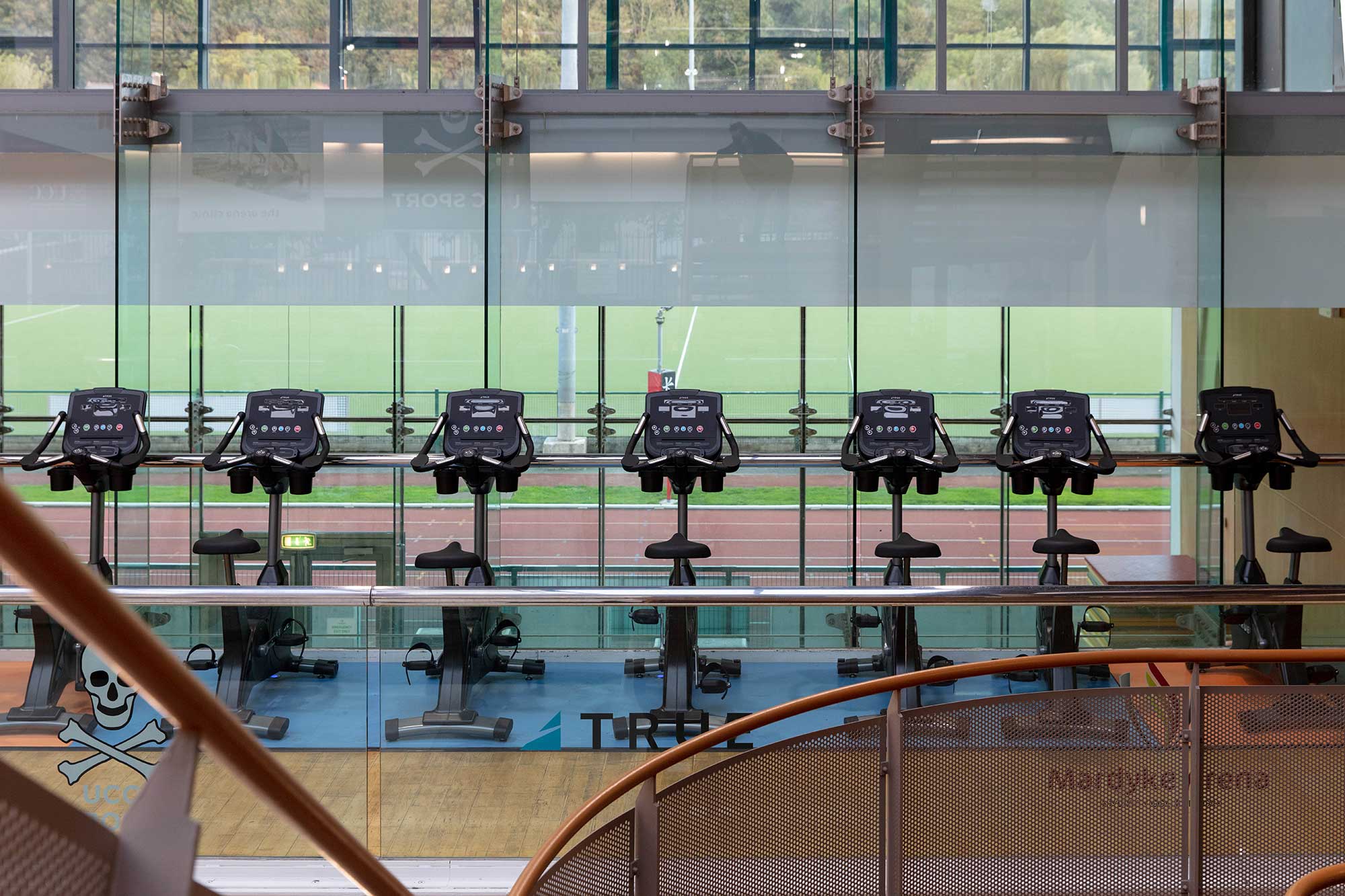 A row of stationary exercise bikes faces large glass windows overlooking a green sports field, with natural light filling the modern gym space.