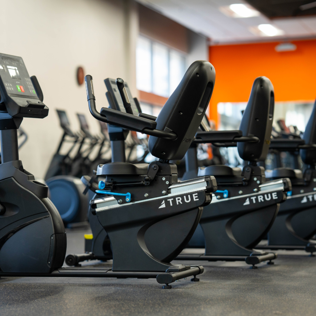 Row of recumbent exercise bikes in a gym with black seats and handles, facing digital screens. Bright orange accent wall and large windows in the background. The gym appears clean and modern.