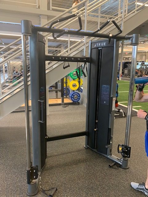 A cable crossover machine at a gym, with adjustable pulleys and handles, stands on a rubber floor beneath a staircase. Weight plates and other gym equipment are visible in the background.